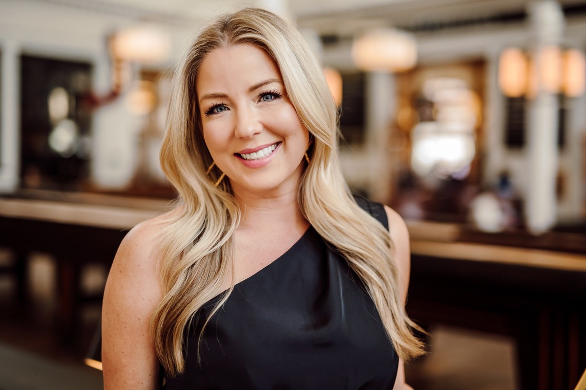 Client with long blonde waves in a black one-shoulder top, smiling in a warm interior