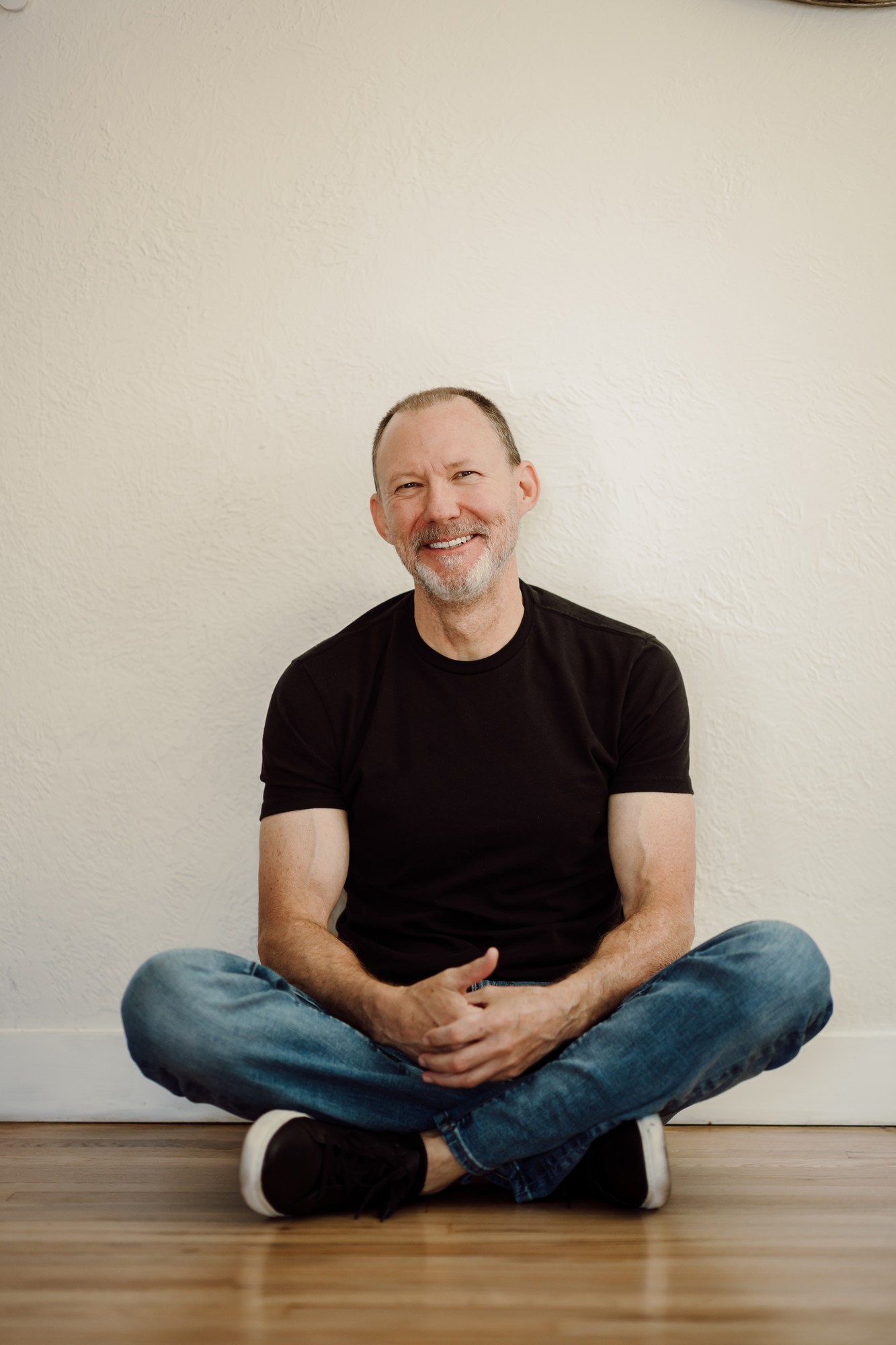 Headshot client in a black t-shirt sitting cross-legged against a white wall